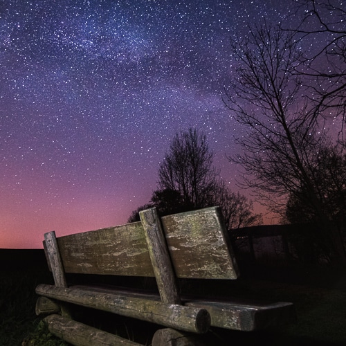 Eine Bank steht in der Nacht auf einem Feld. Im Hintergrund ist ein Himmel mit Milchstrasse zu sehen. Das Foto entstand bei einem Fotoworkshop zur Nachtfotografie in Hohenfelden bei Erfurt.