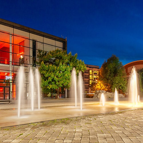 Vor dem erleuchteten Opernhaus in Erfurt sind in der Nacht Springbrunnen aktiv und angeleuchtet. Das Foto entstand bei einem Fotoworkshop Nachtfotografie.